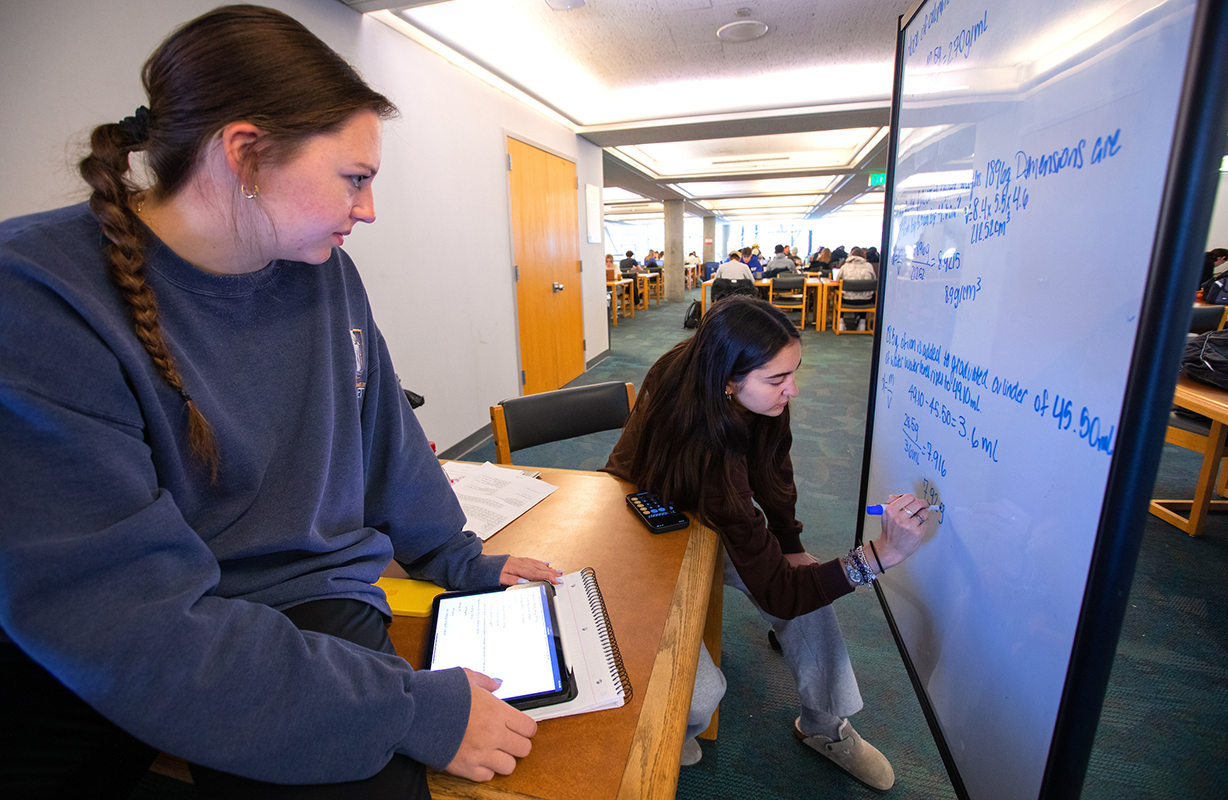 Two female students work out chemistry problems on a whiteboard