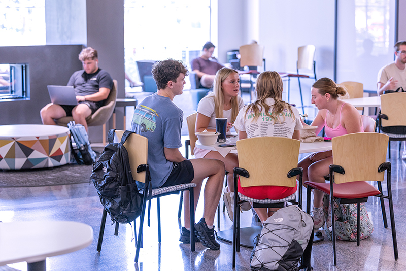 One male and three female students converse at a table in lounge