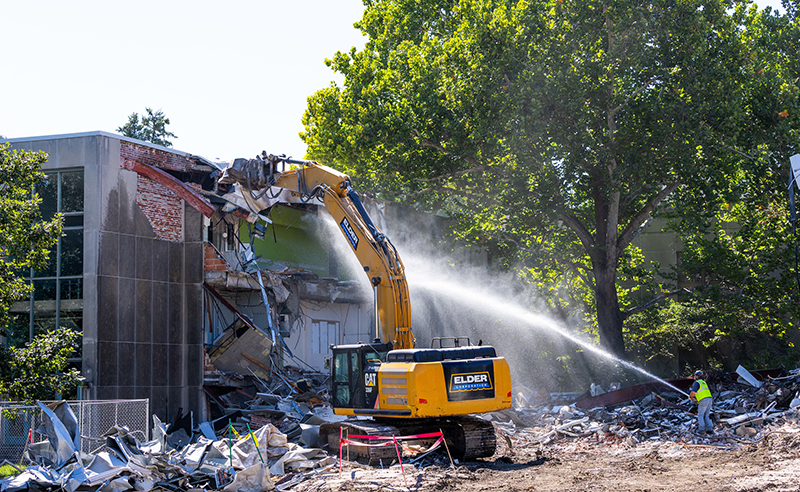 Excavator pulls building wall down, worker hoses down dust