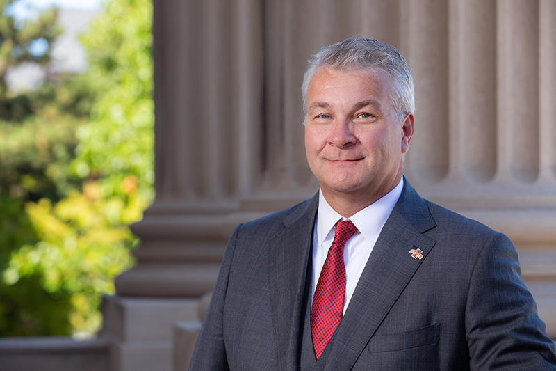 White man in blue suit, red tie next to column