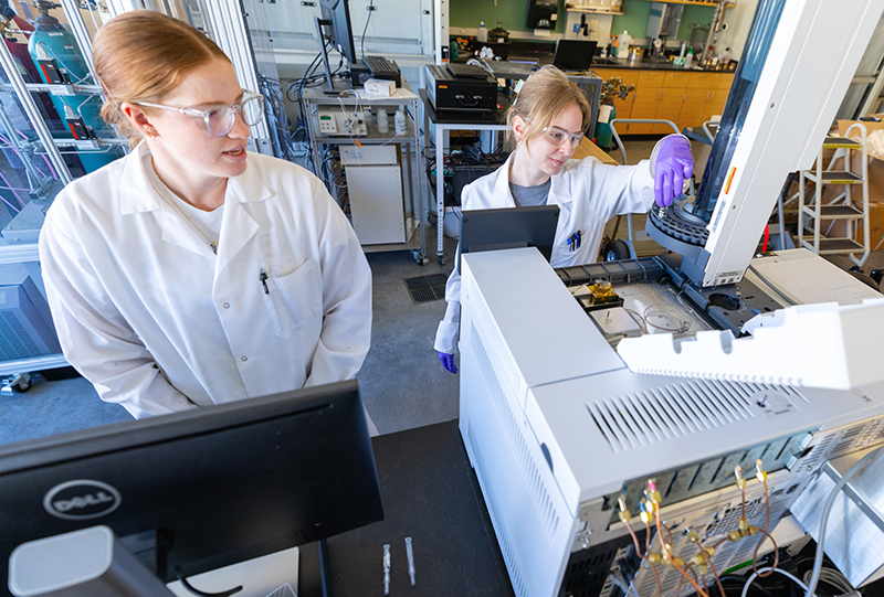 Two female students in white lab coats use chromatography equipm