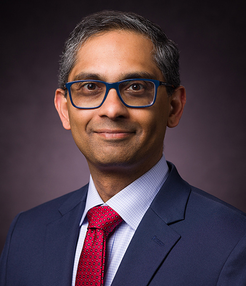 head shot of brown-skinned man in blue suit and blue eyeglasses