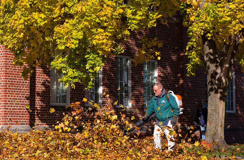 Man with vacuum back pack blows leaves near brick building