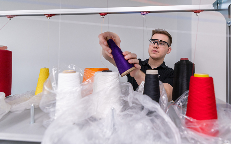 Male wearing protective glasses loads spools on industrial knitt