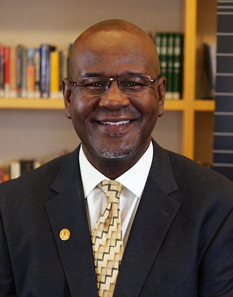 head shot of black man wearing glasses and brown suit with yello