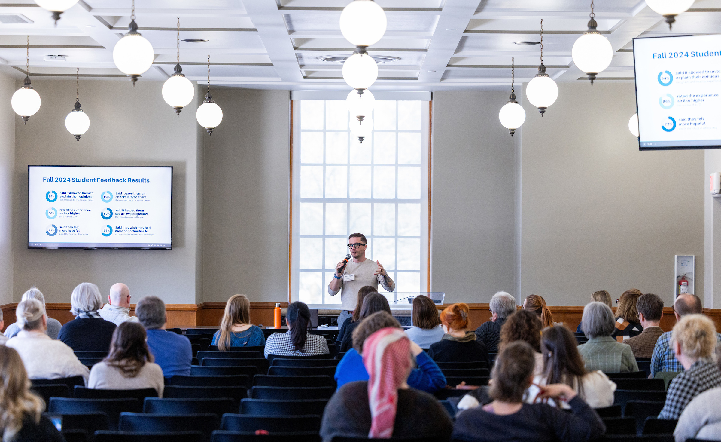 White man in glasses speaks to a seated audience