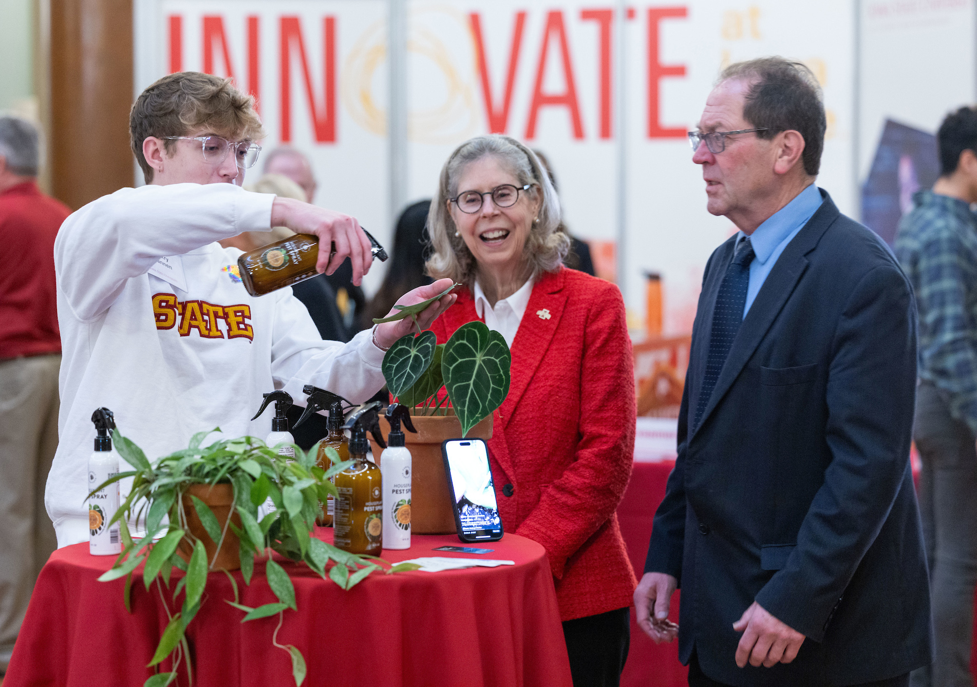 male student spritzes house plant while woman in red blazer and 